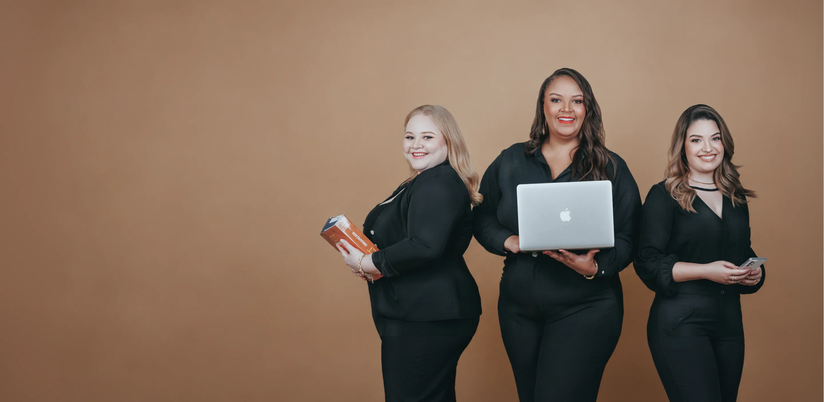 Equipe de advogadas vestidas de preto, sorrindo com notebook, livro e celular, prontas para oferecer assessoria jurídica no escritório de advocacia
